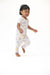A smiling toddler running barefoot in a white short-sleeve pajama set with a pastel multicolor dog pattern, captured against a seamless white studio background.