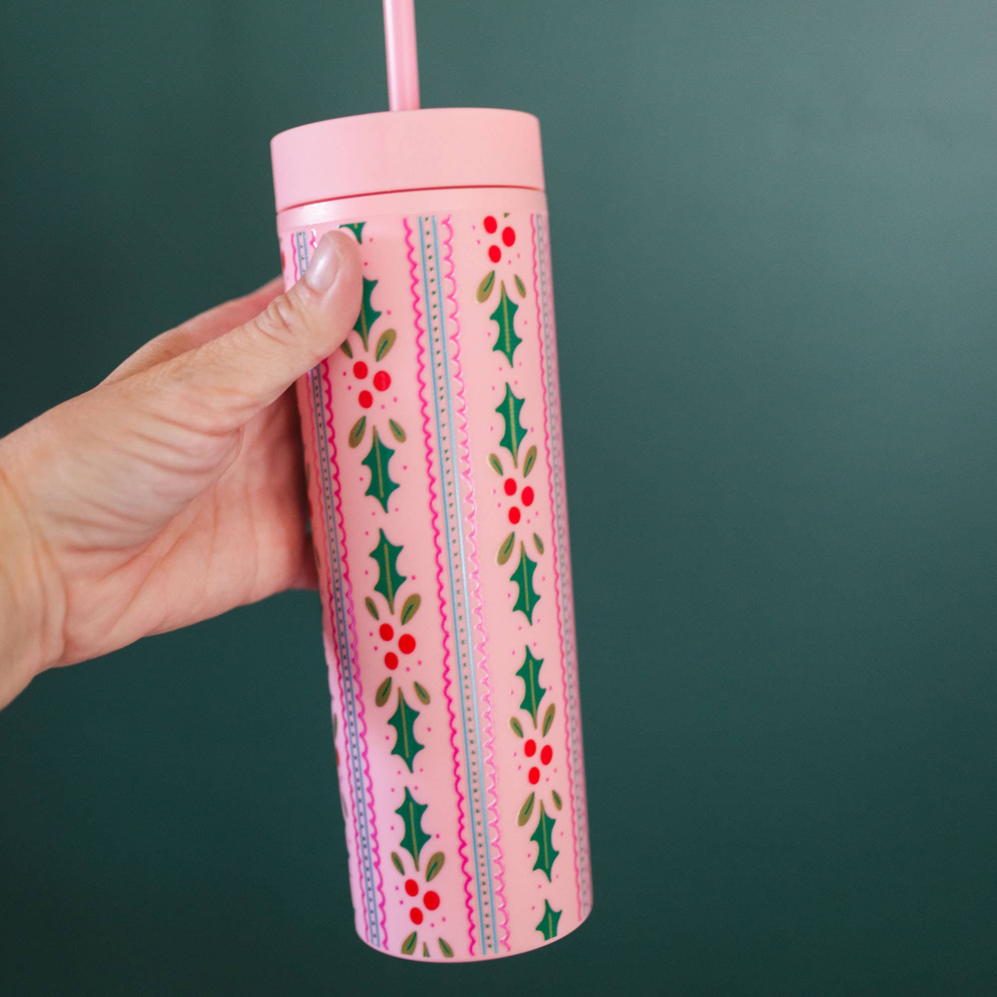 Pink 16 ounce skinny acrylic tumbler with straw held by hand and decorated with holly leaves red berries and scalloped stripes from Pippi Post