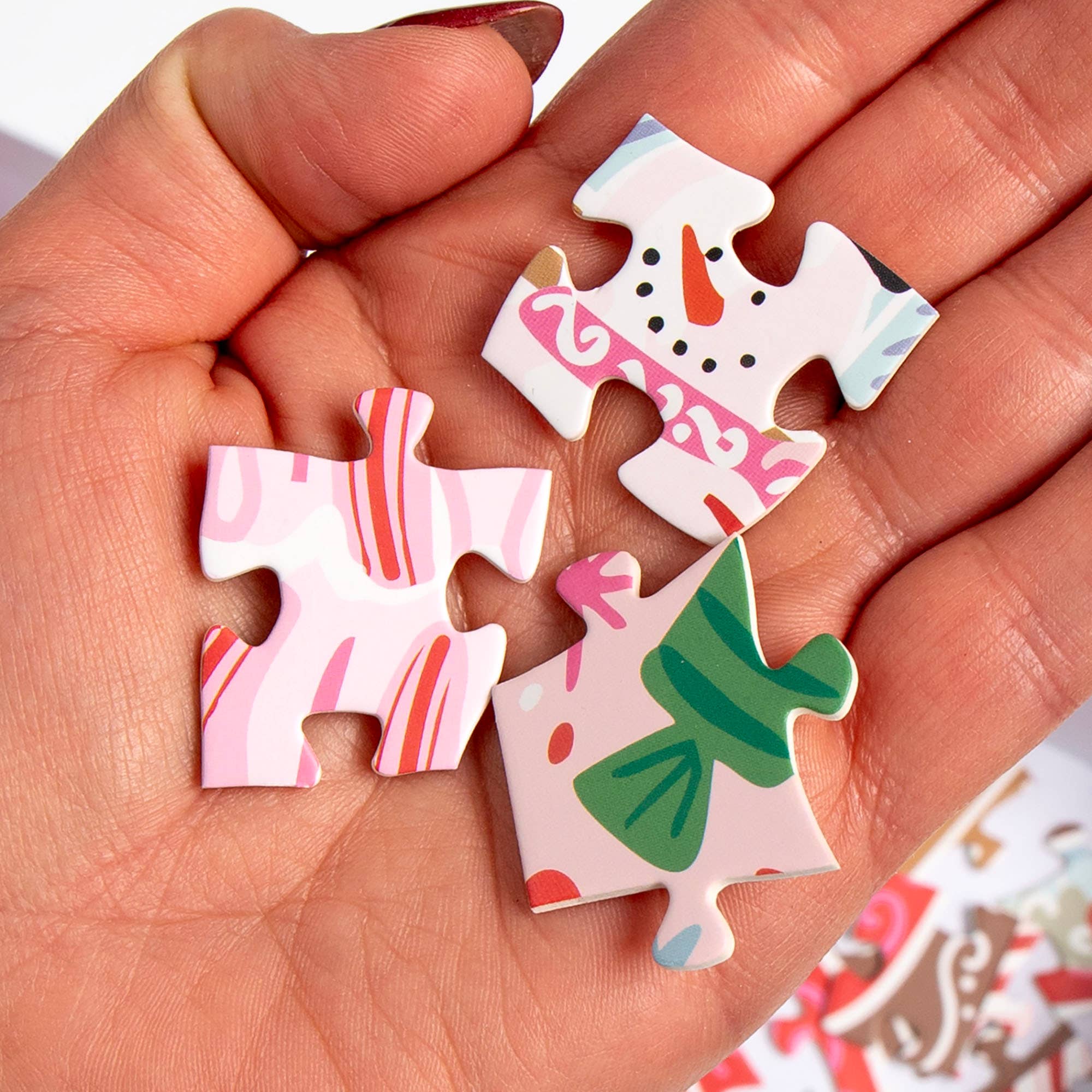 Three colorful pieces from a 500 piece Christmas jigsaw puzzle held in a hand featuring festive gingerbread village artwork by Pippi Post