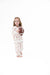 A young girl stands barefoot against a white background, wearing a white long-sleeve two piece set with a football pattern and holding a brown football.