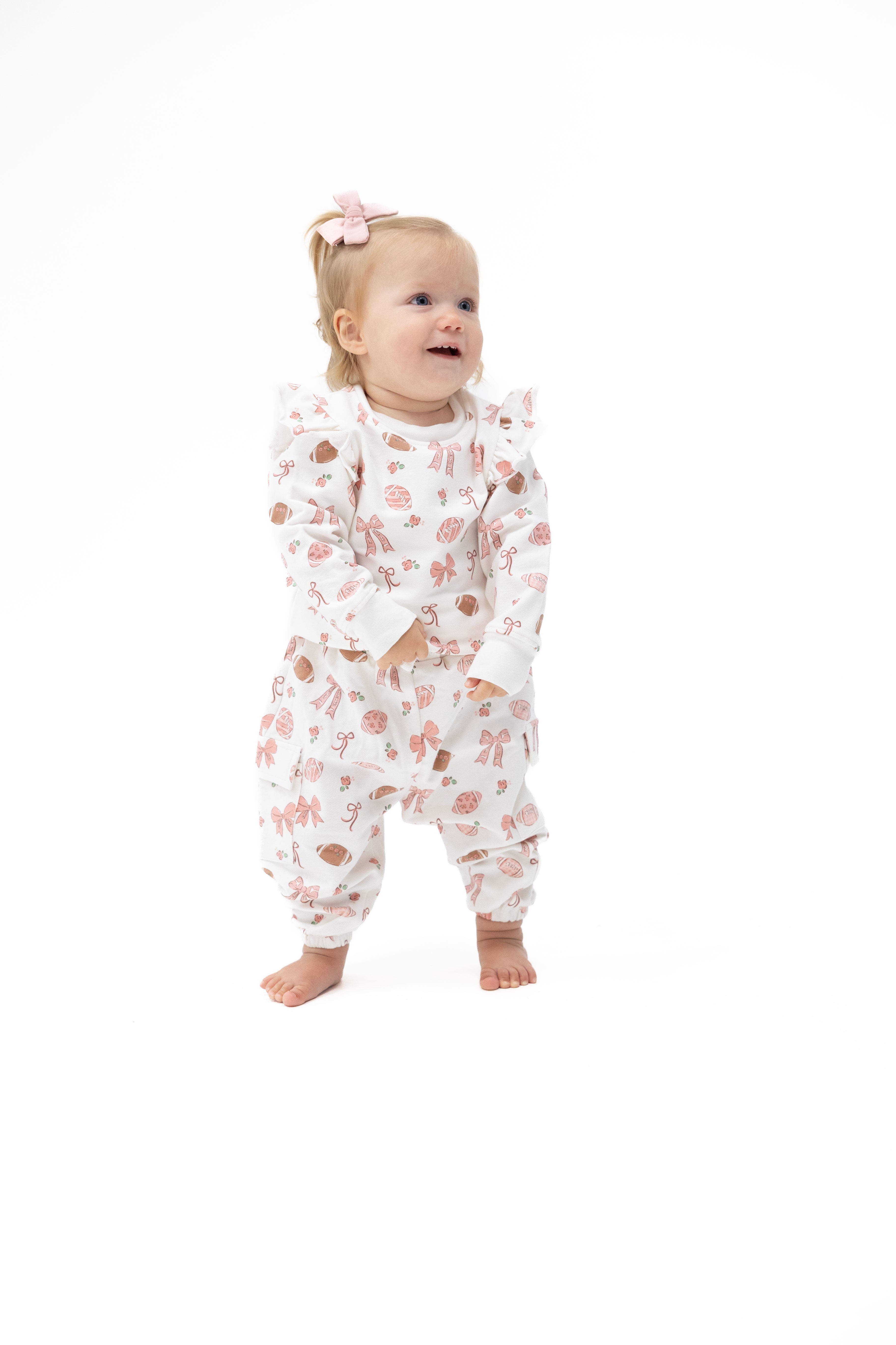 A baby girl wearing a two piece outfit with a pink and brown football print, standing barefoot against a solid white background.