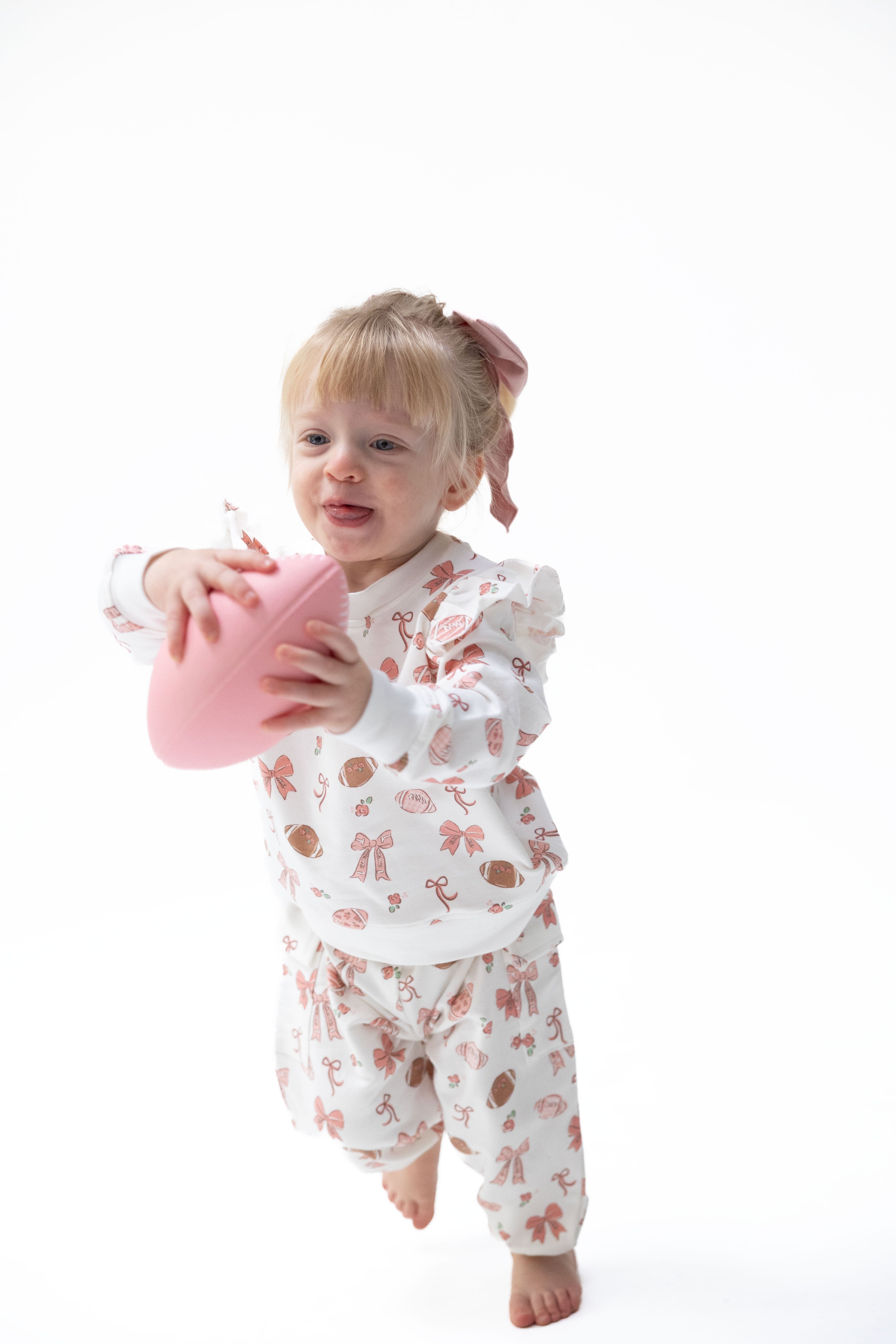 A toddler girl with blonde hair in a pink bow is wearing a white long-sleeve pajama set with a pink and brown football and bow pattern, holding a pink football against a solid white background.