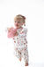 A toddler girl with blonde hair in a pink bow is wearing a white long-sleeve pajama set with a pink and brown football and bow pattern, holding a pink football against a solid white background.