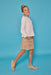 A young girl modeling a white long-sleeve blouse and a beige floral-print mini skirt, paired with beige knit flats, set against a solid blue studio background.