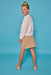 A young girl with blonde hair in a bun wearing a white knit long-sleeve top and a beige floral-print mini skirt, posing against a solid blue studio background.