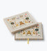 Illustrated keepsake baby book by Rifle Paper Co. with woodland animals and floral details on the cover plus a yellow ribbon bookmark beneath a second book