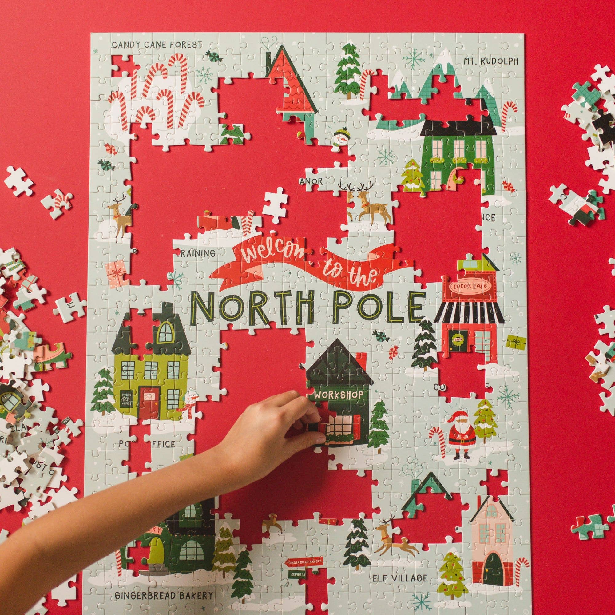 In progress Welcome to the North Pole 500 piece jigsaw puzzle showing Santa village on a red background with loose pieces nearby and a hand placing a piece from Pippi Post