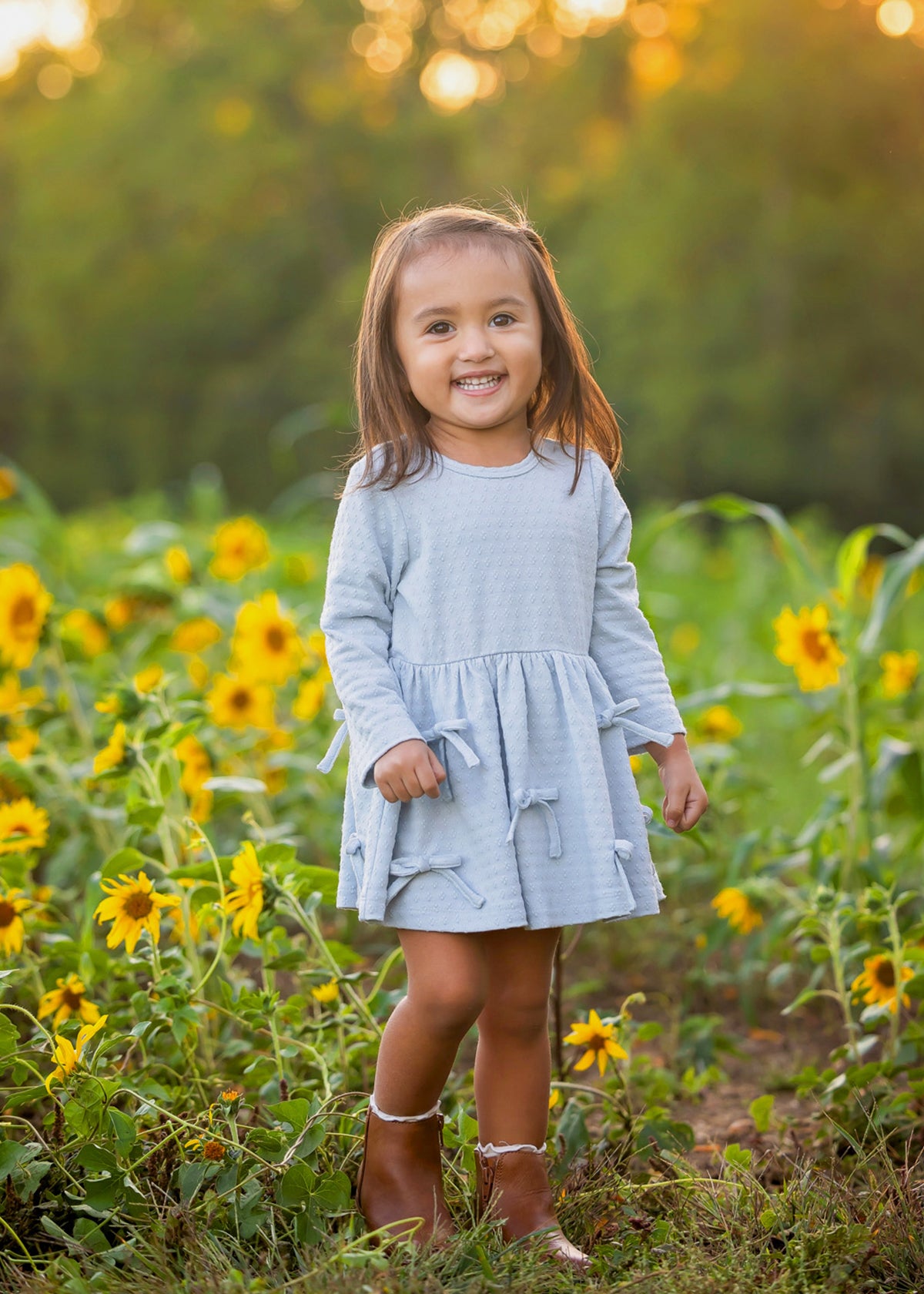 A young girl smiling in a sunflower field, wearing a light blue long-sleeve tiered dress and brown boots.