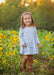 A young girl smiling in a sunflower field, wearing a light blue long-sleeve tiered dress and brown boots.
