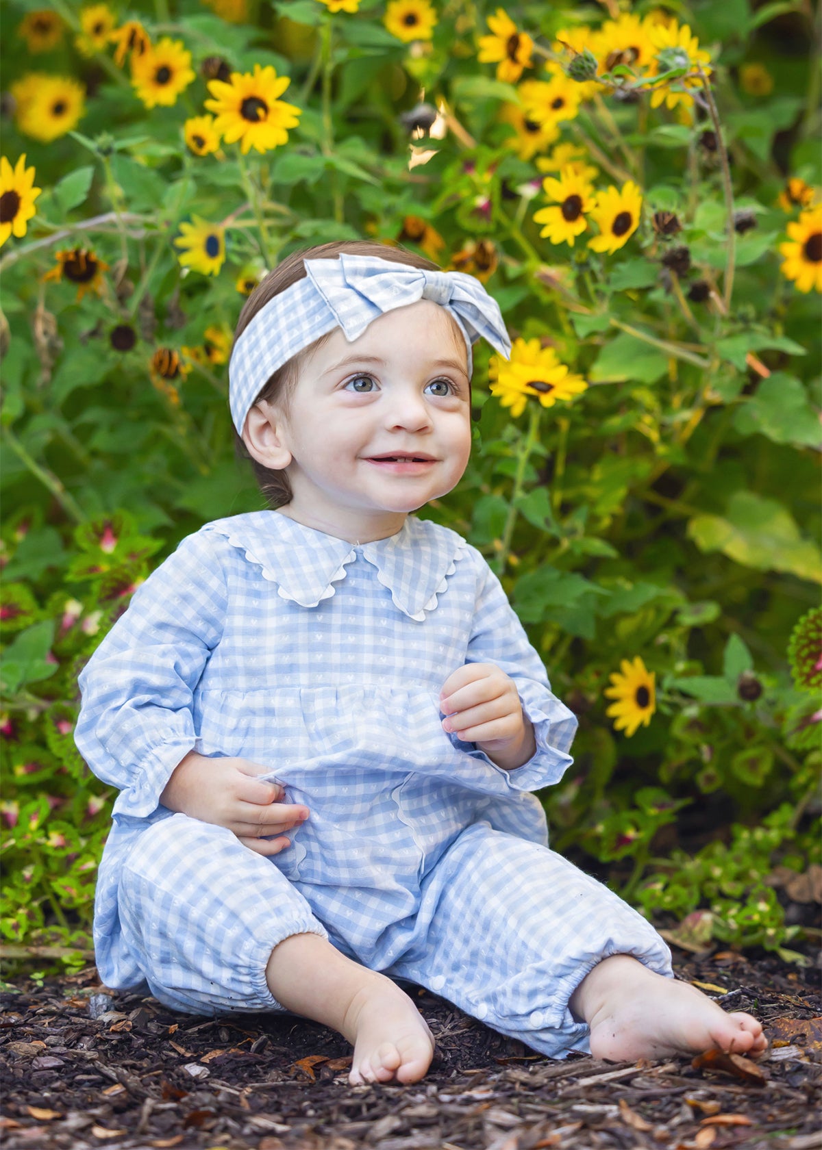 A baby girl wearing a light blue and white gingham romper and matching headband sits on a bed of mulch in a garden with yellow sunflowers in the background.