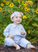 A baby girl wearing a light blue and white gingham romper and matching headband sits on a bed of mulch in a garden with yellow sunflowers in the background.