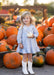 A young girl stands in a pumpkin patch holding a small white pumpkin. She is wearing a light blue and white gingham dress with a white cowboy boot on each foot.