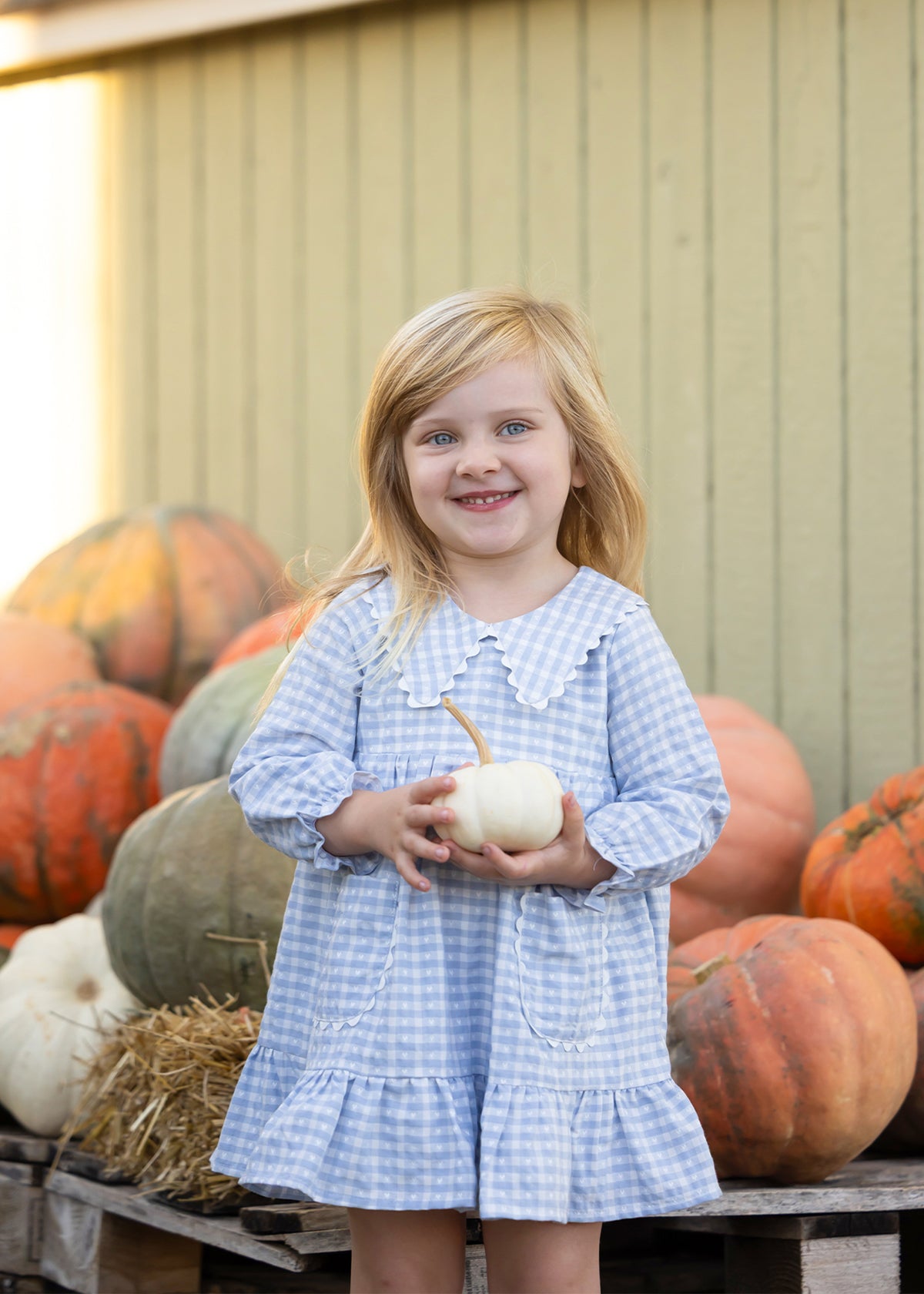 A young girl with blonde hair stands smiling in front of a pile of pumpkins, wearing a light blue and white gingham dress and holding a small white pumpkin.