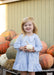A young girl with blonde hair stands smiling in front of a pile of pumpkins, wearing a light blue and white gingham dress and holding a small white pumpkin.