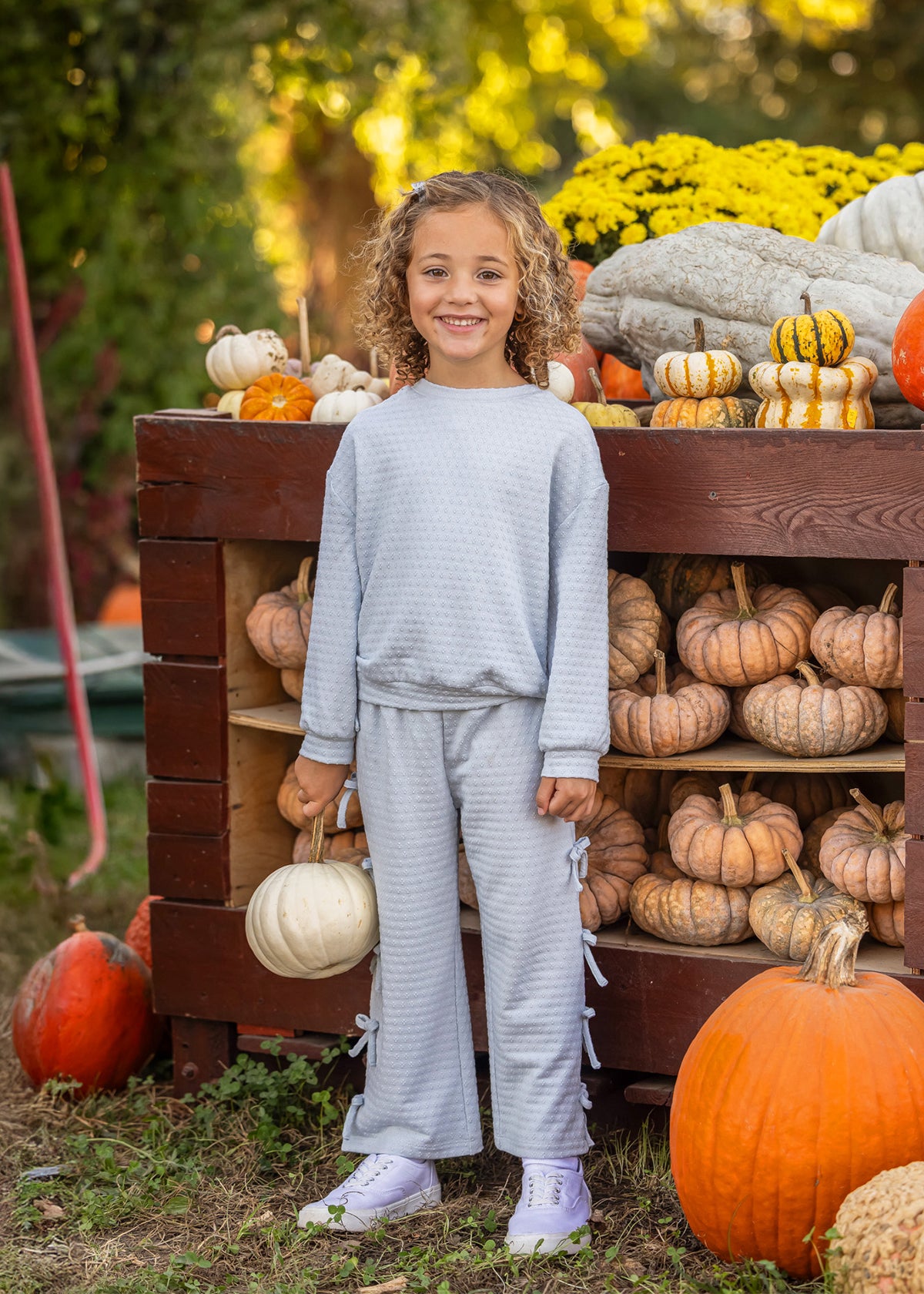 A young girl stands outdoors in front of a wooden crate filled with pumpkins, wearing a light blue-gray waffle-knit long-sleeve top and matching wide-leg pants with side tie details, paired with white sneakers.