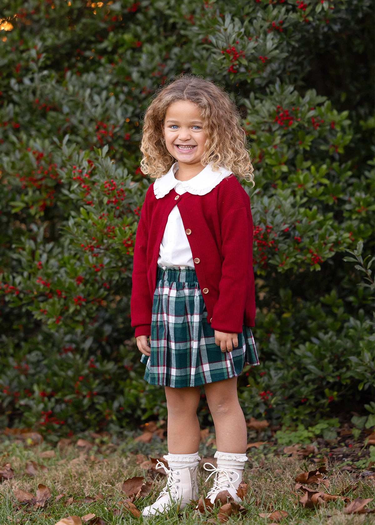 A young girl with curly light brown hair stands outdoors in front of a leafy green hedge with red berries. She is wearing a festive school holiday outfit consisting of a deep red cardigan over a white collared shirt, a green and white plaid skirt, and white lace-up boots.