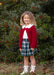 A young girl with curly light brown hair stands outdoors in front of a leafy green hedge with red berries. She is wearing a festive school holiday outfit consisting of a deep red cardigan over a white collared shirt, a green and white plaid skirt, and white lace-up boots.