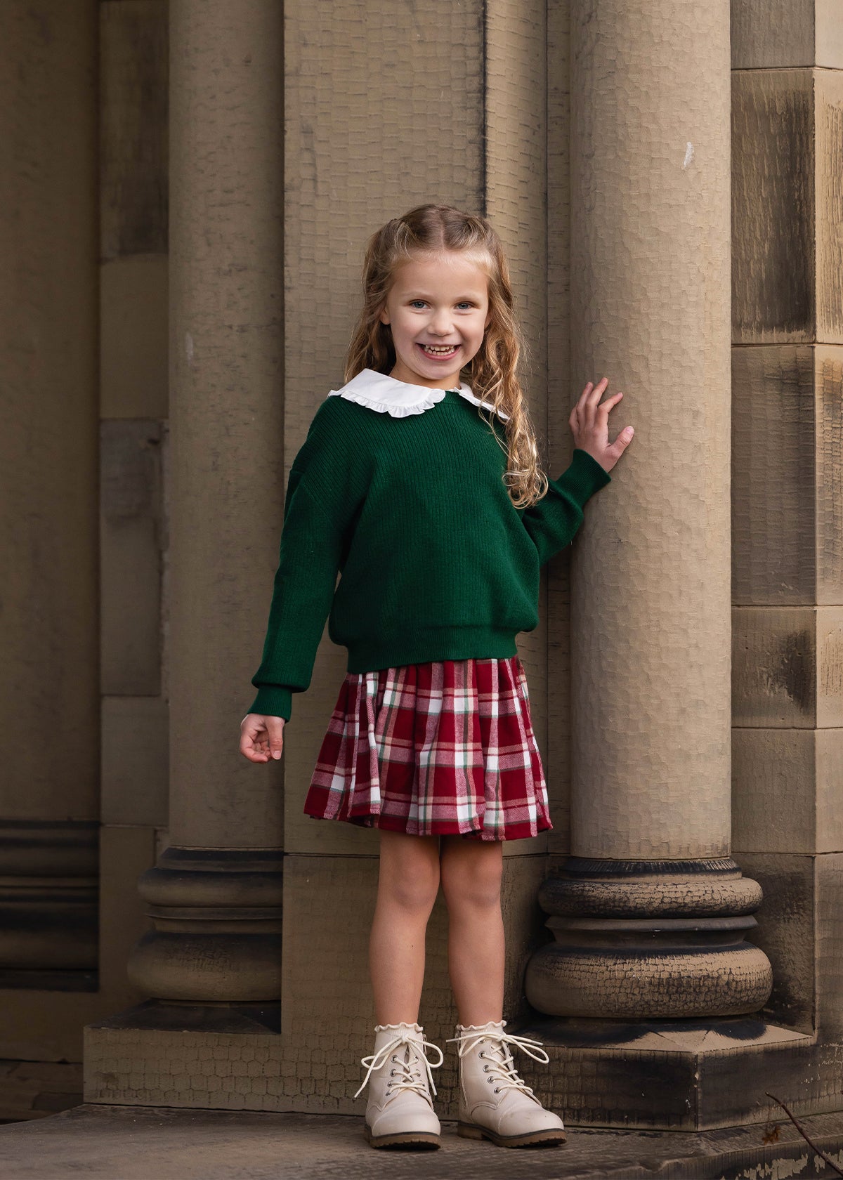 A young girl with blonde hair stands in front of large stone columns, wearing a dark green crewneck sweater with a white Peter Pan collar, a red and white plaid pleated skirt, and beige lace-up boots.