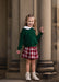 A young girl with blonde hair stands in front of large stone columns, wearing a dark green crewneck sweater with a white Peter Pan collar, a red and white plaid pleated skirt, and beige lace-up boots.