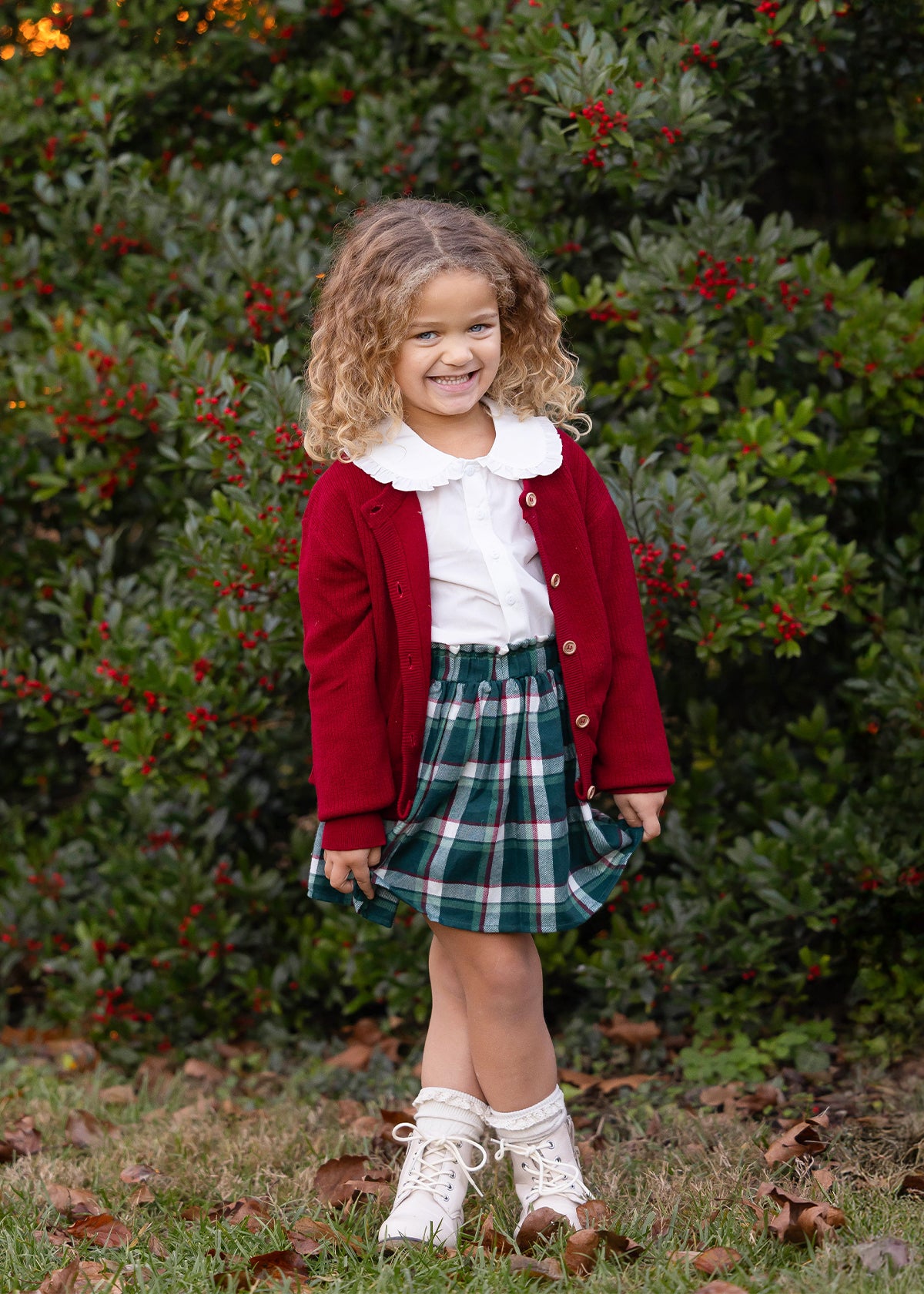 A young girl with curly light brown hair stands outdoors in front of a green leafy bush with red berries. She is wearing a dark red cardigan over a white collared shirt, a green and white plaid skirt, and white lace-up ankle boots.