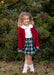 A young girl with curly light brown hair stands outdoors in front of a green leafy bush with red berries. She is wearing a dark red cardigan over a white collared shirt, a green and white plaid skirt, and white lace-up ankle boots.