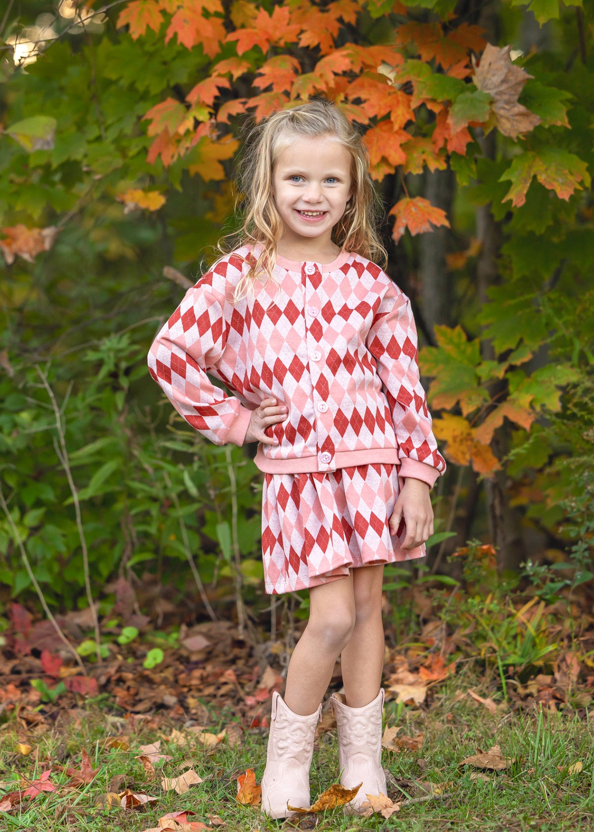 A young girl stands outdoors in a wooded setting with autumn foliage, wearing a pink and red argyle patterned long-sleeve top and matching shorts, paired with light pink boots.