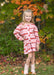 A young girl stands outdoors in a wooded setting with autumn foliage, wearing a pink and red argyle patterned long-sleeve top and matching shorts, paired with light pink boots.