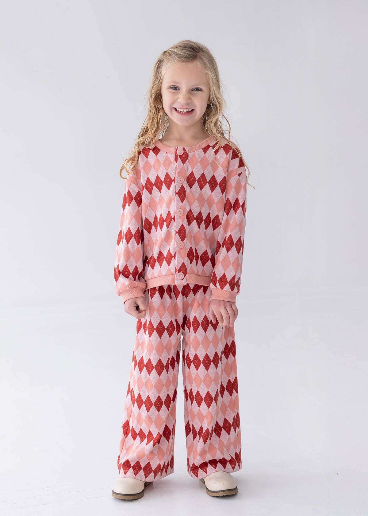 A young girl with blonde hair stands against a light gray studio background wearing a matching two-piece pajama set with a red and pink argyle pattern.