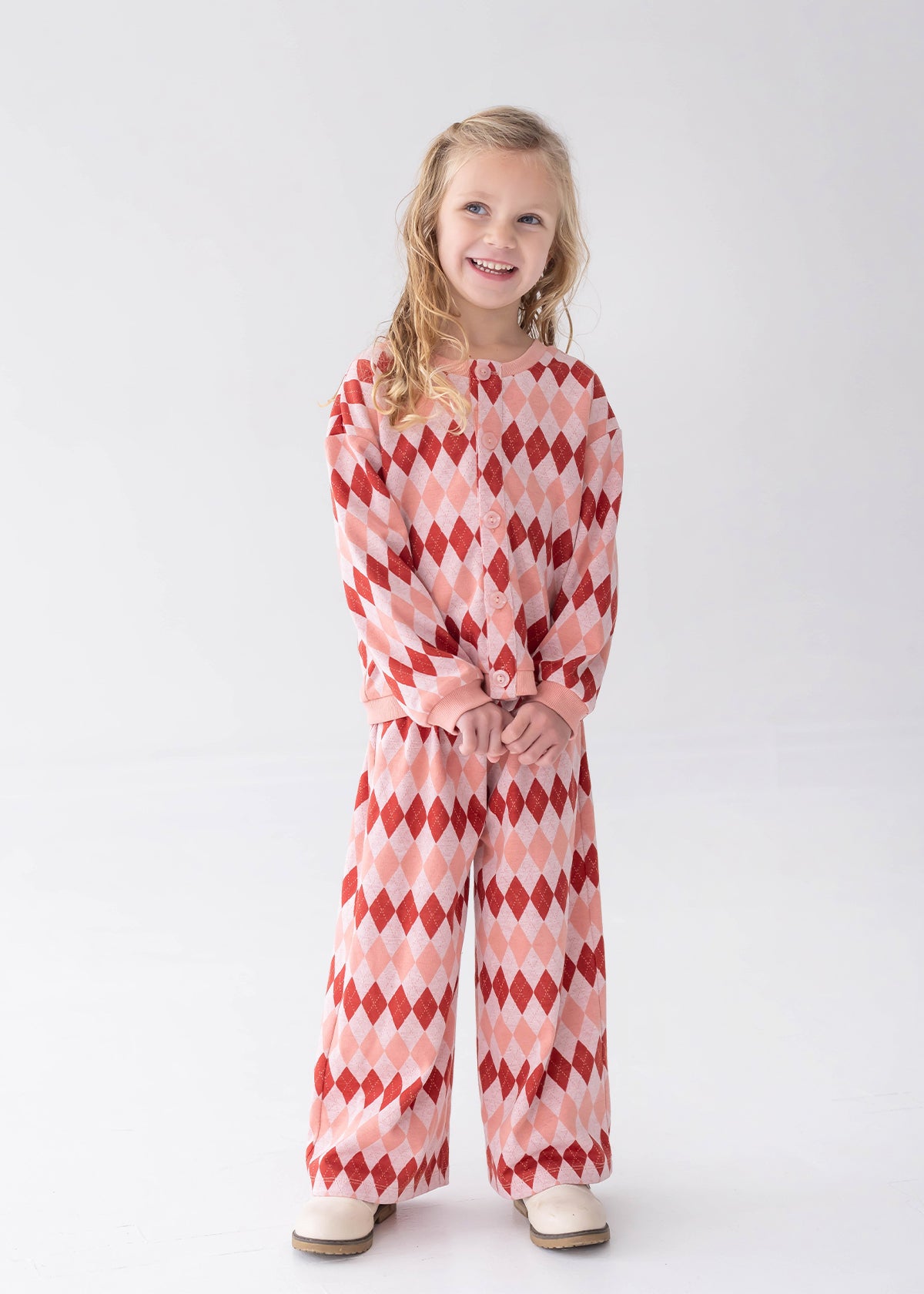 A young girl with blonde hair stands against a light gray studio background wearing a matching two-piece lounge set with a red and pink harlequin diamond pattern. The set includes a long-sleeve top and wide-leg pants.