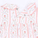 A close-up of a baby dress featuring a delicate floral and bow pattern in pink, green, and lavender on a white background, with scalloped edges and snap buttons.