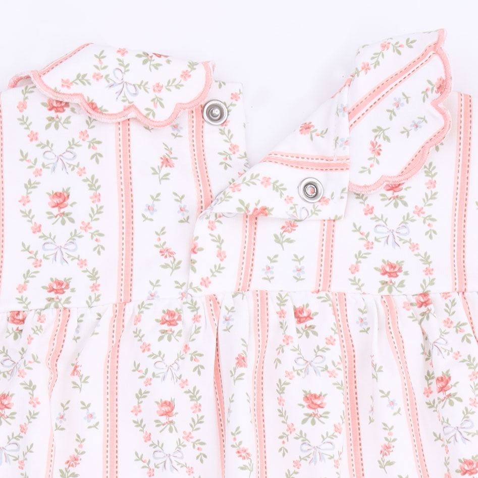 A close-up of a baby dress featuring a delicate floral and bow pattern in pink, green, and lavender on a white background, with scalloped edges and snap buttons.