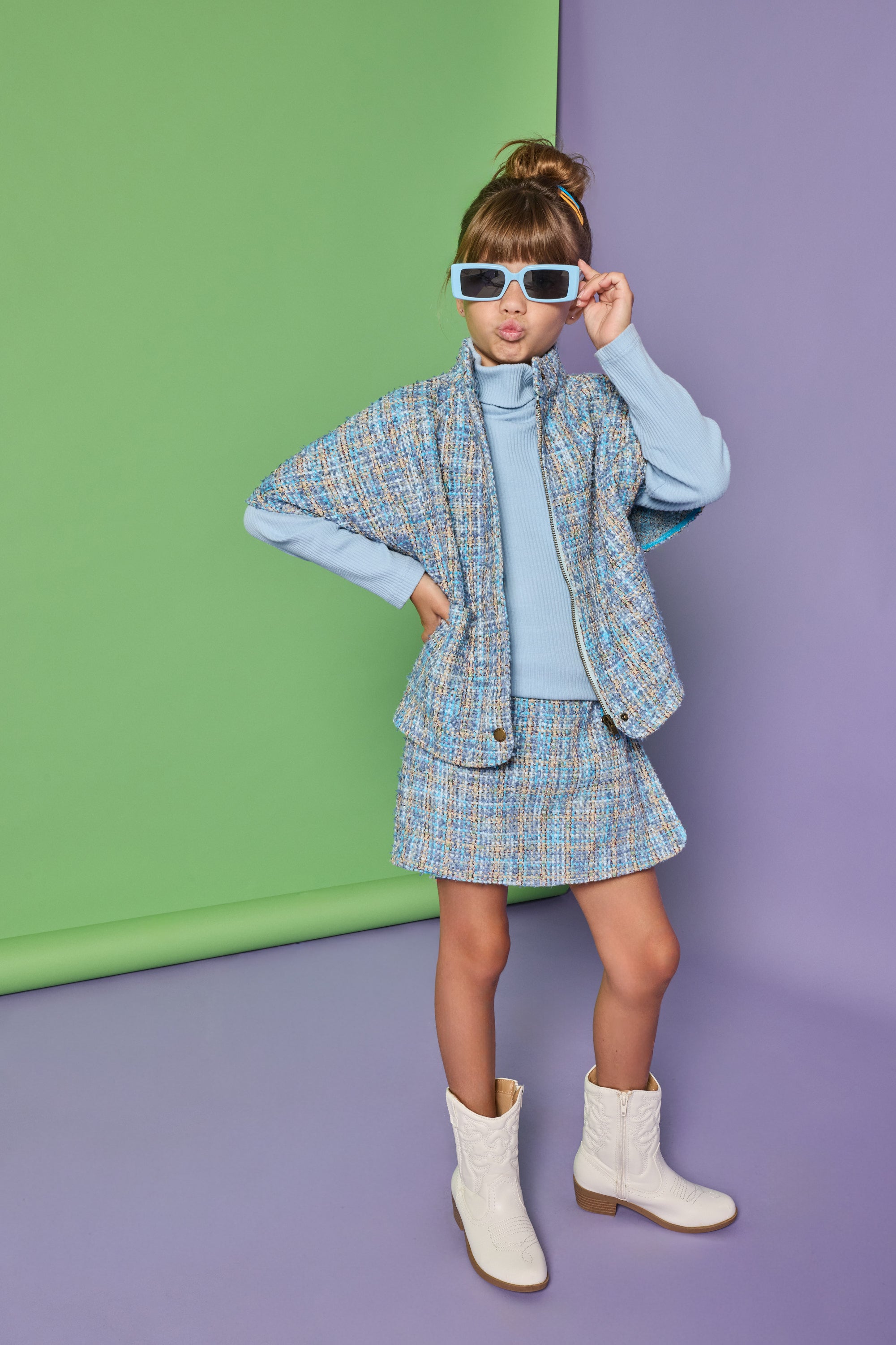 A young girl models a light blue and white tweed cape and matching mini skirt set, styled with a light blue mock neck top, white western-style boots, and white-framed sunglasses, standing against a split green and purple studio backdrop.