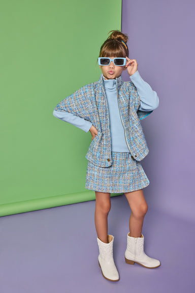 A young girl models a light blue and white tweed cape and matching mini skirt set, styled with a light blue mock neck top, white western-style boots, and white-framed sunglasses, standing against a split green and purple studio backdrop.