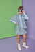 A young girl models a light blue and white tweed cape and matching mini skirt set, styled with a light blue mock neck top, white western-style boots, and white-framed sunglasses, standing against a split green and purple studio backdrop.