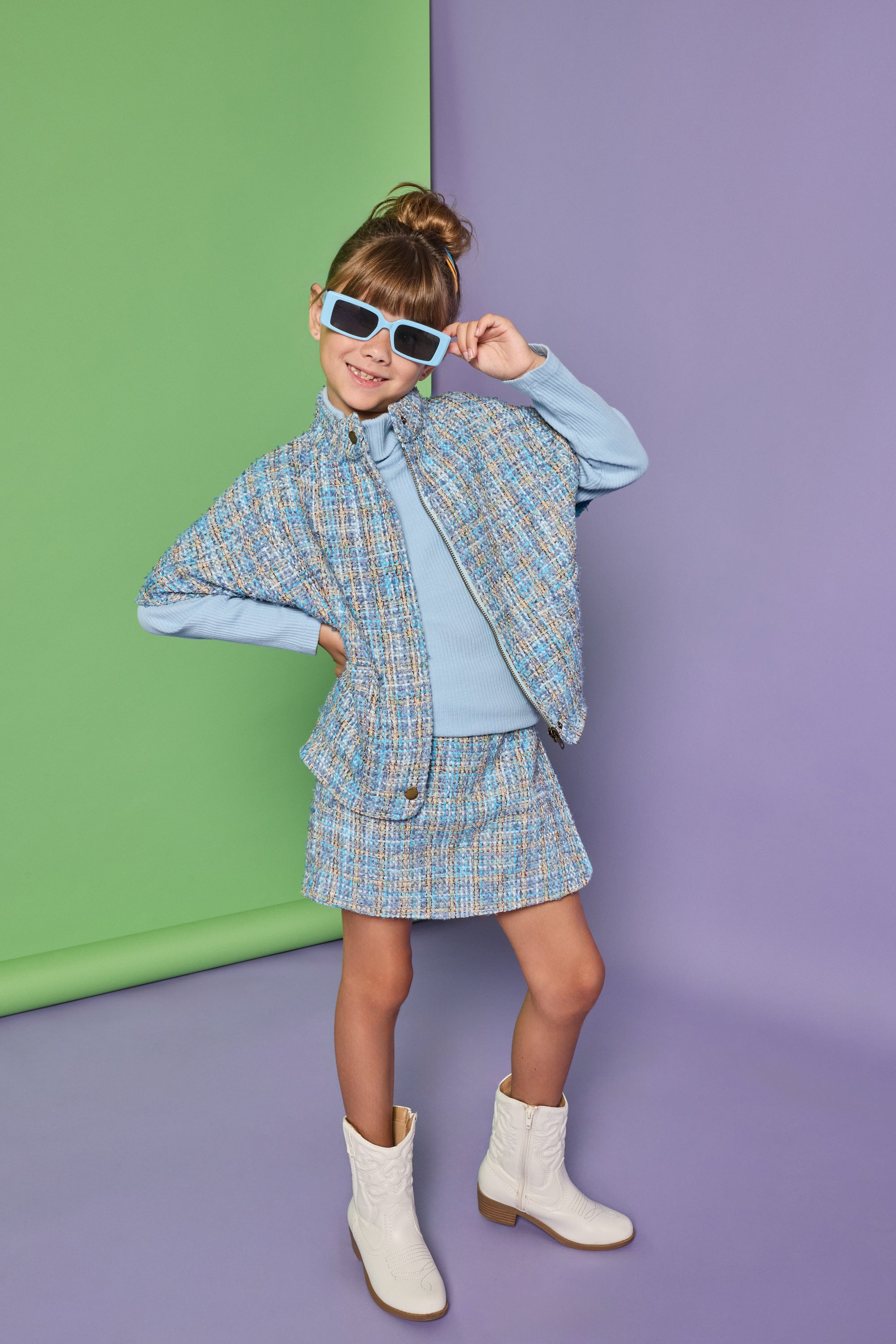 A young girl models a light blue tweed cape and matching mini skirt set with a light blue top and white ankle boots, posing against a split green and purple studio backdrop.