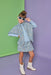 A young girl models a light blue tweed cape and matching mini skirt set with a light blue top and white ankle boots, posing against a split green and purple studio backdrop.