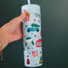 Pippi Post Christmas movie themed skinny tumbler with straw featuring popcorn car holiday sign ice skates and colorful seasonal icons held in one hand