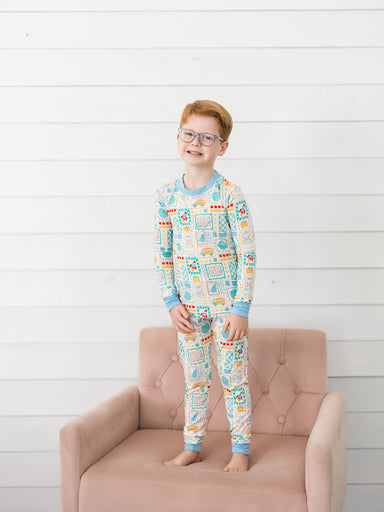 A young boy with red hair and glasses stands barefoot on a dusty pink tufted armchair, wearing a long-sleeve pajama set with a colorful school themed pattern on a white background.