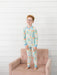 A young boy with red hair and glasses stands barefoot on a dusty pink tufted armchair, wearing a long-sleeve pajama set with a colorful school themed pattern on a white background.
