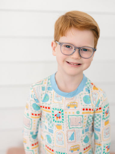 A young boy with red hair and glasses wearing a long-sleeve crewneck shirt with a colorful school-themed pattern, set against a white paneled wall background.