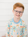 A young boy with red hair and glasses wearing a long-sleeve crewneck shirt with a colorful school-themed pattern, set against a white paneled wall background.
