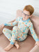 A young boy with red hair and glasses sits on a dusty pink armchair, wearing a long-sleeve pajama set with a colorful school themed pattern on a cream background.