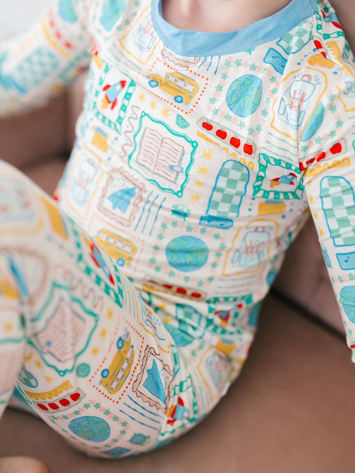 A close-up of a child wearing a long-sleeve one-piece pajama with a colorful school-themed print on a cream background, featuring blue, yellow, and green illustrations of cars, books, and buildings.