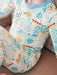A close-up of a child wearing a long-sleeve one-piece pajama with a colorful school-themed print on a cream background, featuring blue, yellow, and green illustrations of cars, books, and buildings.
