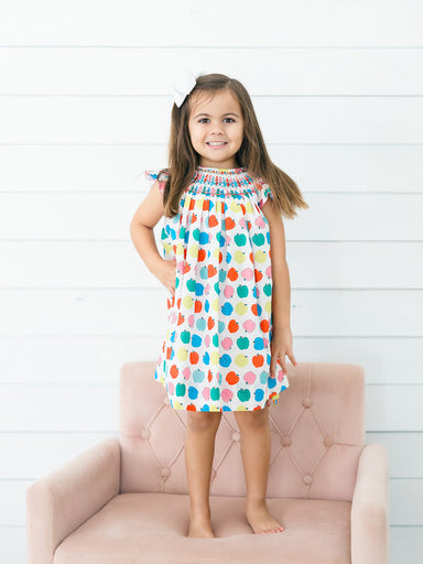 A young girl stands barefoot on a pink tufted chair against a white shiplap wall, wearing a sleeveless smocked dress with a colorful polka-dot pattern.