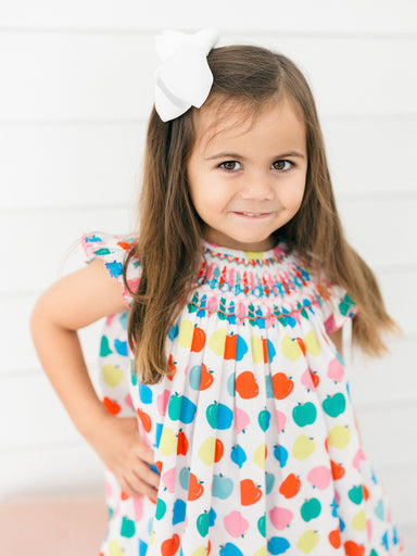 A young girl with long brown hair wearing a white dress with a colorful apple print and a white bow in her hair, posing against a white paneled wall.