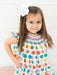 A young girl with long brown hair wearing a white dress with a colorful apple print and a white bow in her hair, posing against a white paneled wall.