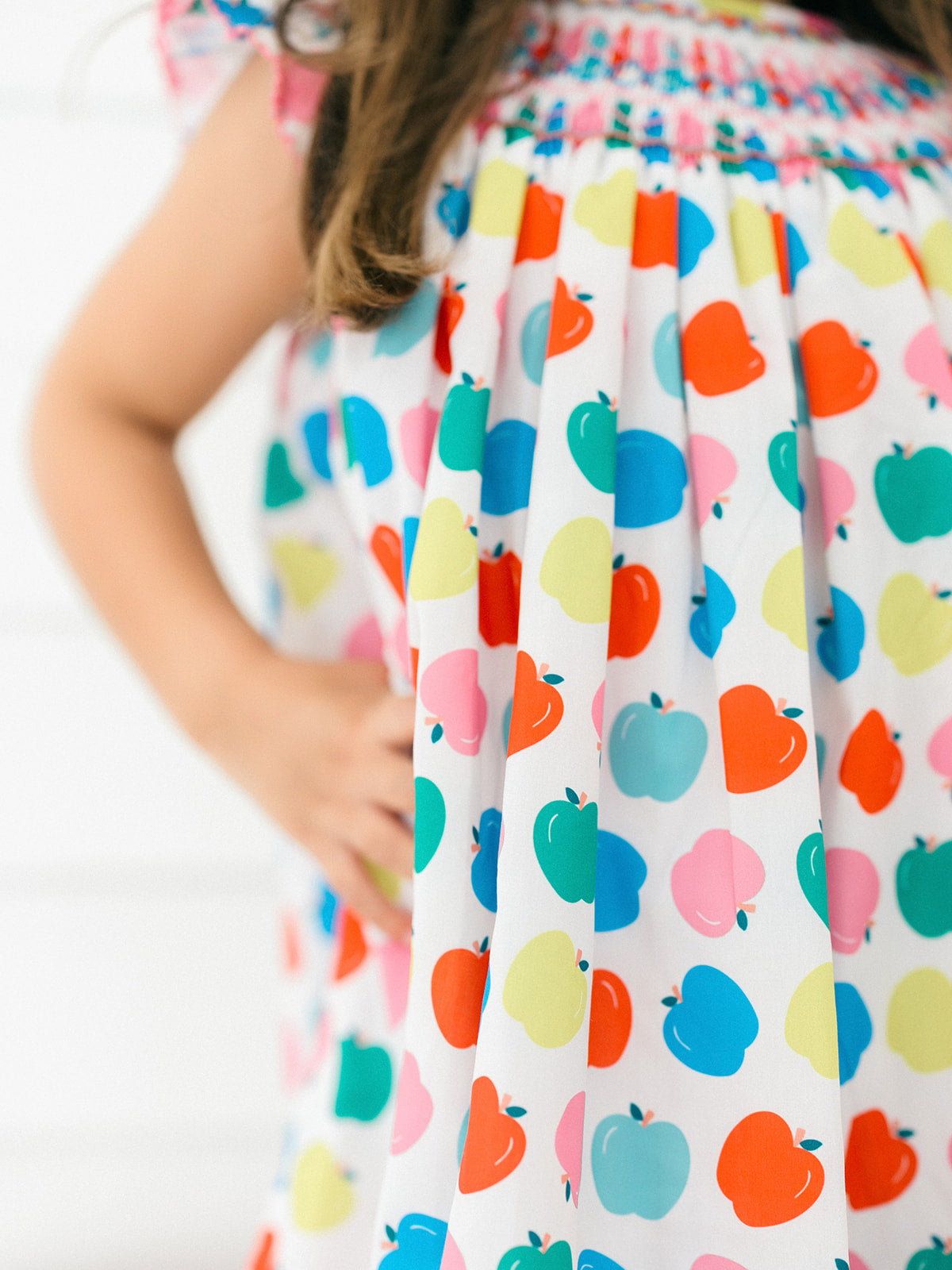 A close-up of a child wearing a white dress with a colorful pattern of stylized apples in blue, green, orange, pink, and yellow. The dress has a smocked bodice and is shown against a bright white background.