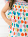 A close-up of a child wearing a white dress with a colorful pattern of stylized apples in blue, green, orange, pink, and yellow. The dress has a smocked bodice and is shown against a bright white background.