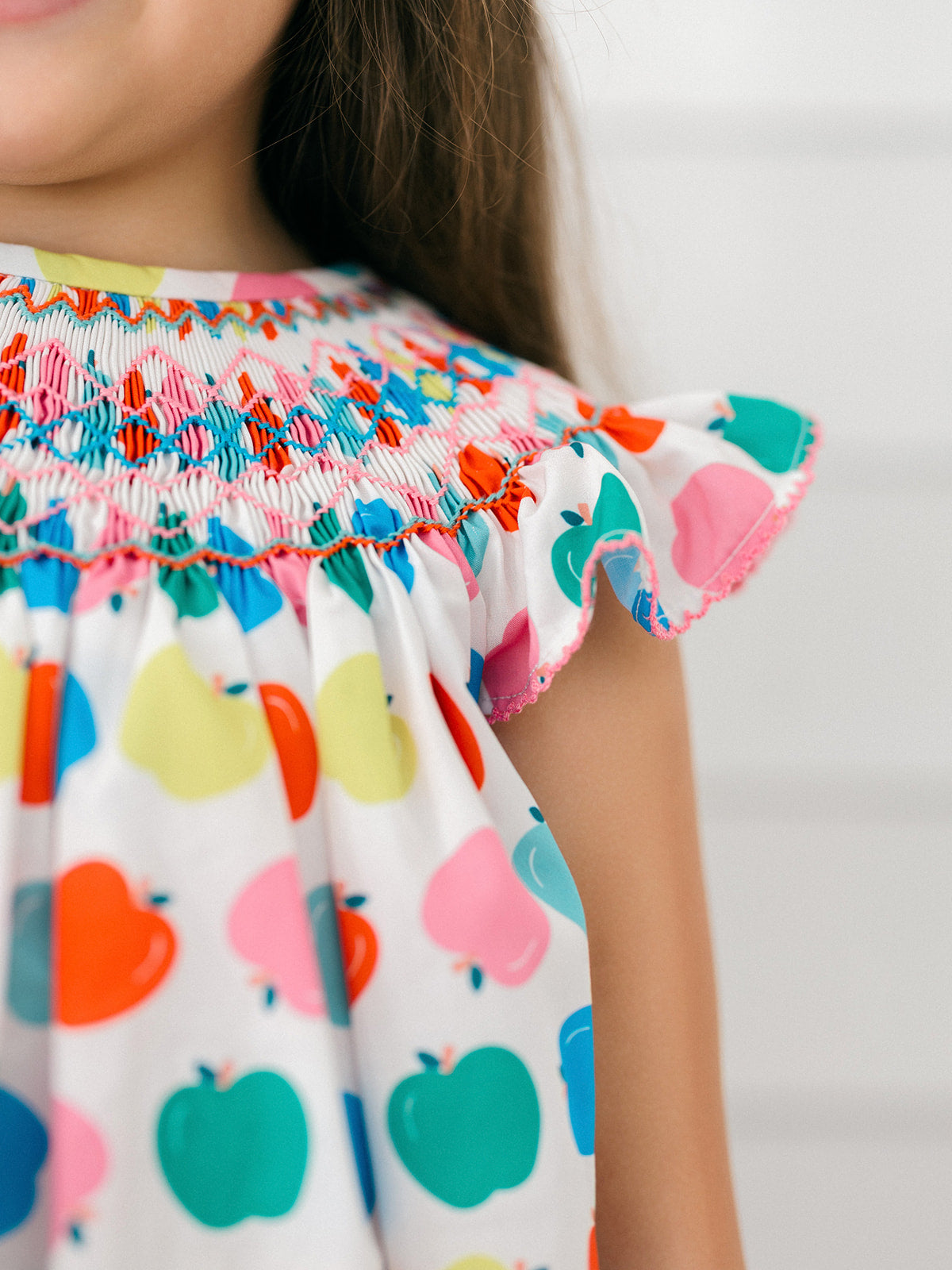 Close-up of a child wearing a white smocked dress with colorful apple print and ruffled sleeves.